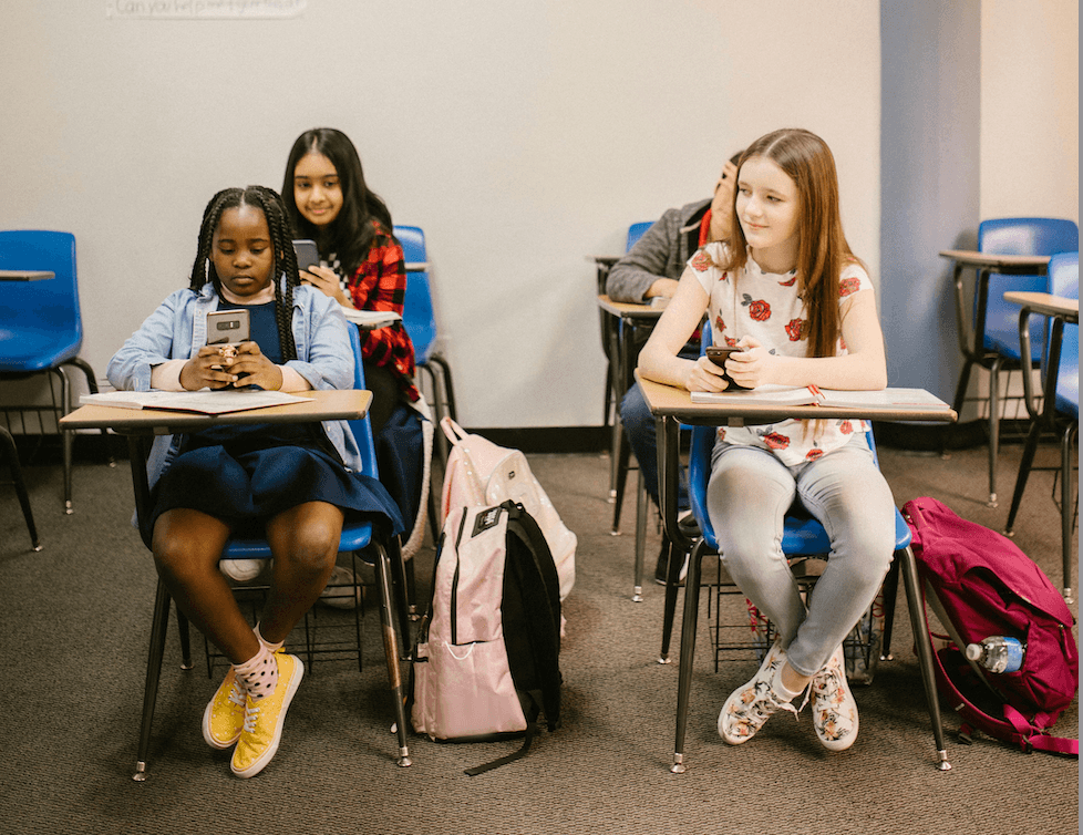 Four students sit at desks in a classroom. Two girls in the front row look at their phones, while two students in the back row interact and smile. Backpacks are on the floor beside their desks.